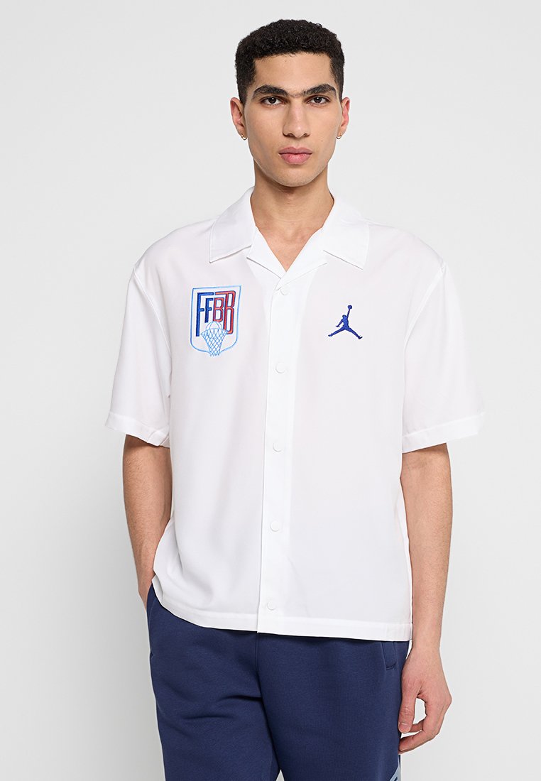 Camisa de manga corta blanca con botones que presenta bordados temáticos de baloncesto en azul, incluyendo un logo y detalles en la zona del pecho. Tejido suave.