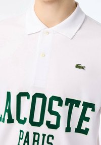 White polo shirt with green "LACOSTE PARIS" text and a small embroidered green crocodile logo on the chest.