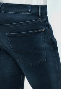 Κοντινό πλάνο πίσω τσέπης σκούρου μπλε τζιν με ετικέτα "for all mankind" και διακριτική ραφή σε εφαρμοστά παντελόνια.