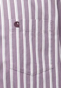 Rövidujjú ing világoslila színben, fehér függőleges csíkokkal. Különlegessége egy foltvarrott logóval ellátott zseb a mellrészen. Pamut anyag.