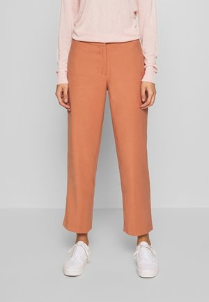 Personne portant un pantalon droit couleur pêche, un haut à manches longues rose clair, et des baskets blanches, debout contre un mur gris.