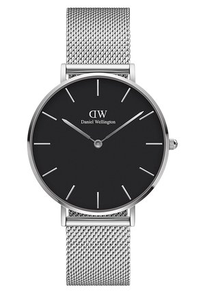 Poignet avec montre en maille argentée et cadran noir, reposant dans la poche d'un pantalon à carreaux gris, portant un haut rose clair à manches longues.