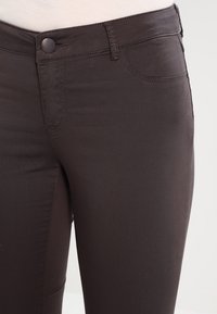 Tmavě hnědé přiléhavé džíny s hladkým povrchem, s předním zapínáním na knoflík, bočními kapsami a jemnými švy.