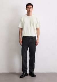 Mężczyzna stoi przy białej ścianie, ubrany w t-shirt z krótkim rękawem w kolorze off-white, czarne dżinsy i czarne buty na sznurowadła, patrząc prosto przed siebie.