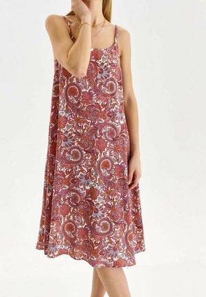 Femme portant une robe sans manches longueur genou avec un motif cachemire multicolore rouge, blanc et bleu, posant avec la main près du visage.