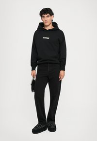 Sudadera negra con el logo "BARROW", pantalones vaqueros negros y zapatillas de patrón negro, sosteniendo una bolsa negra. Conjunto simple y casual.