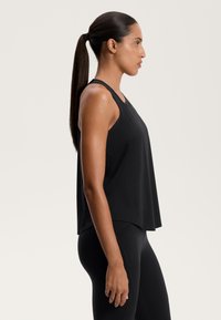 Crna bezrukava majica od glatkog materijala, s opuštenim krojem i zaobljenim rubom, u kombinaciji s crnim tajicama. Bočni prikaz.