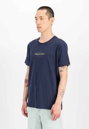 Mann mit kurzen Haaren, der ein marineblaues T-Shirt und helle Jeans trägt, mit Tattoos auf beiden Unterarmen, steht vor einem schlichten weißen Hintergrund.