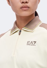 Gros plan d'une personne portant un polo zippé beige et crème avec un col marron et le logo EA7 Emporio Armani sur la poitrine.