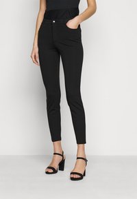 Svarta åtsittande byxor med en slät textur och en elegant design, med sidofickor och en liten silverknapp. Bärs med svarta klacksandaler.