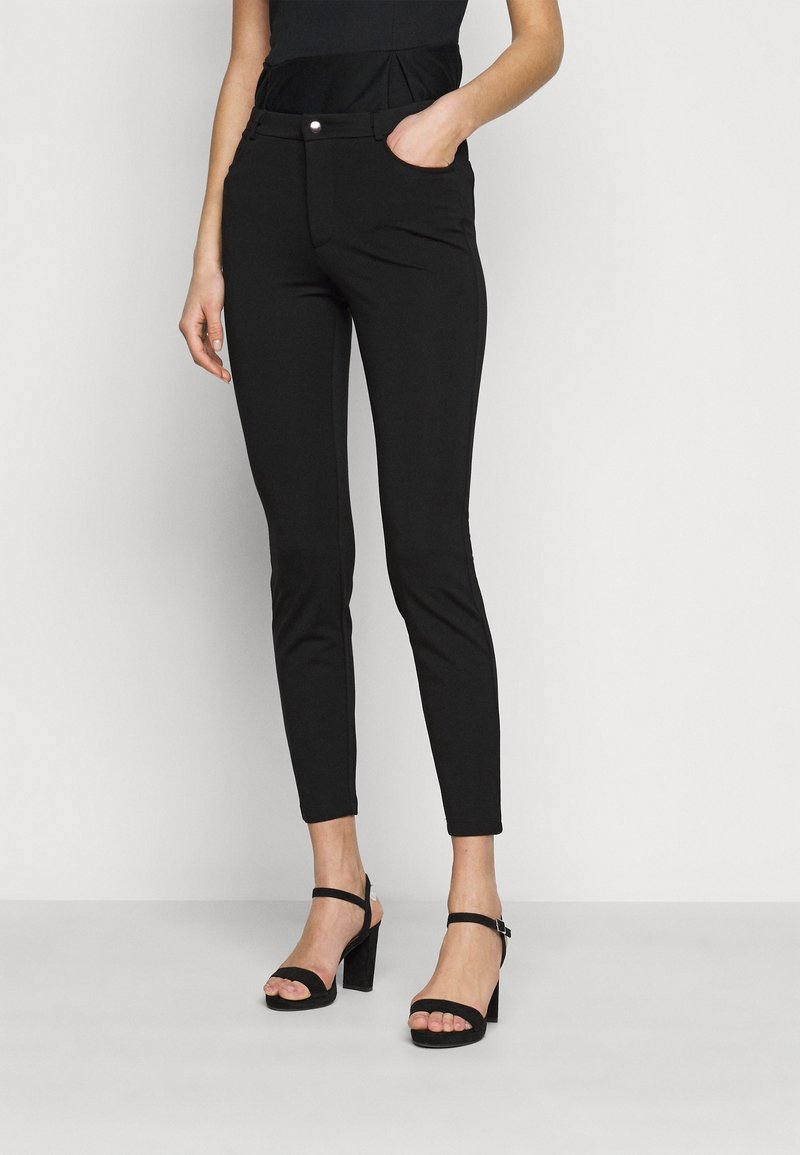 Svarta åtsittande byxor med en slät textur och en elegant design, med sidofickor och en liten silverknapp. Bärs med svarta klacksandaler.