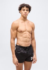 Joven con cabello rizado, vistiendo pantalones cortos de baño negros, de pie contra un fondo claro y liso, mostrando un torso musculoso.