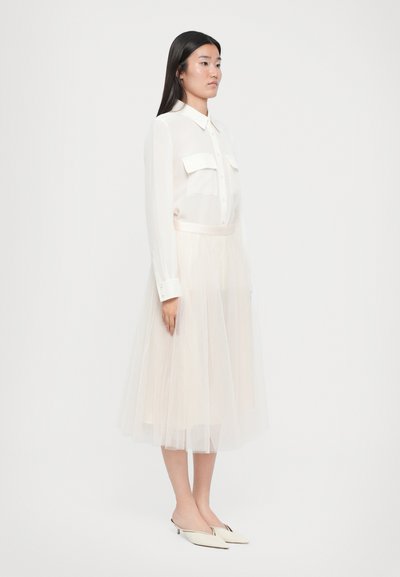 Transparante witte blouse met knoopdetails en twee voorzakken, gecombineerd met een zachte, gelaagde, lichte tule rok en crème puntige schoenen.