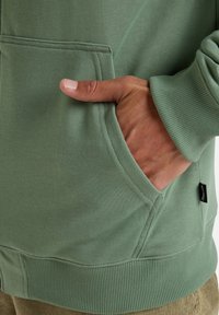 Hoodie vert clair, fabriqué en coton, avec des cuffs et un ourlet côtelés. Il présente une poche kangourou et une petite étiquette noire sur le côté.