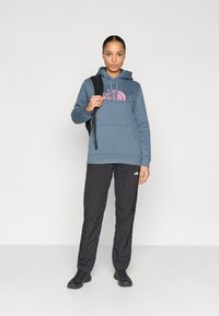 Σκούρα μπλε φούτερ με κουκούλα και ροζ λογότυπο, μαύρο ελαφρύ παντελόνι και μαύρα παπούτσια, το μοντέλο κρατάει ένα μαύρο σακίδιο πλάτης, ουδέτερο φόντο.