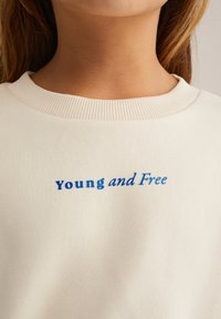 Krakuma pelēka sporta krekls ar ribotām malām, kurā zīmogots "jauns un brīvs" zilo krāsā. Mīksta auduma tekstūra un brīva piegriezums.