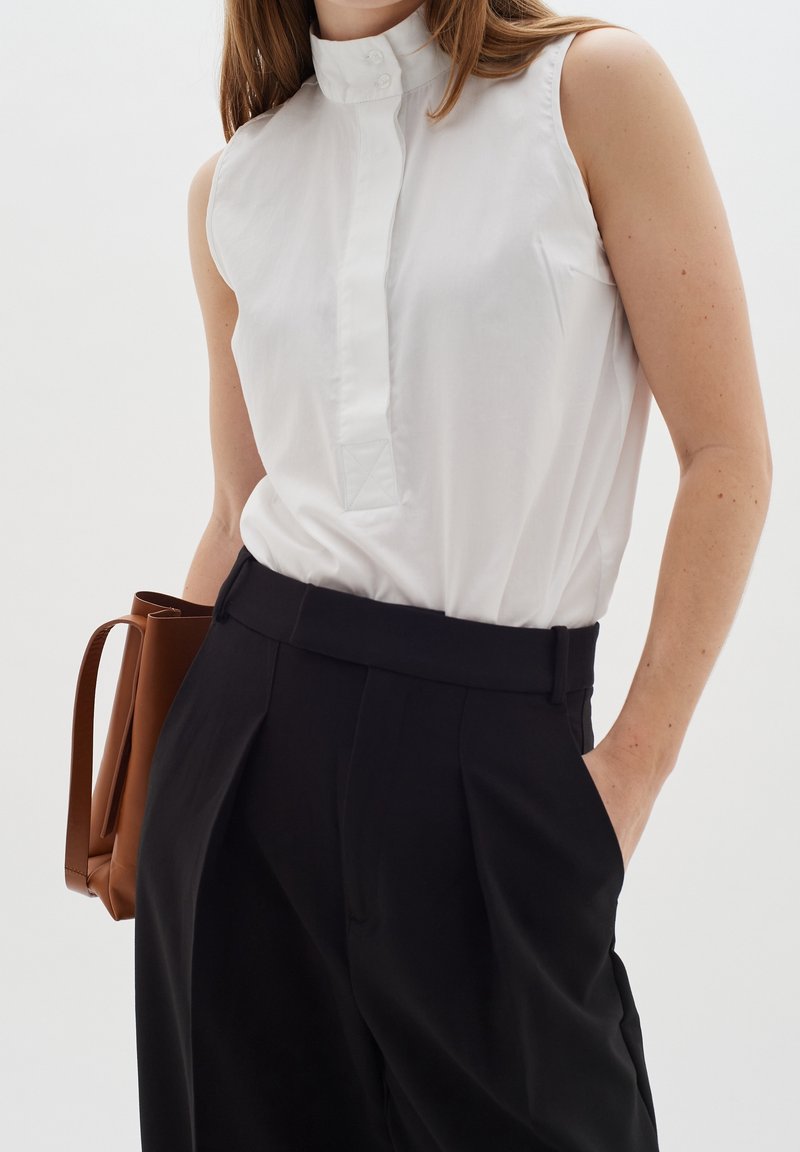 Femme portant un chemisier blanc sans manches boutonné et un pantalon noir taille haute, tenant un sac à main en cuir marron avec une main dans la poche.