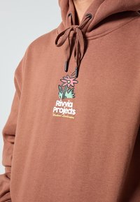 Smeđa dukserica s kapuljačom na vezanje, s grafikom cvijeća i tekstom "Rivvia Projects Weekend Landscapes" u bijeloj i žutoj boji.