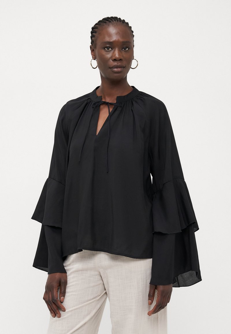 Black, long-sleeve blouse with a tie neck, features layered bell sleeves and a relaxed cut. Worn over light-colored, textured pants.