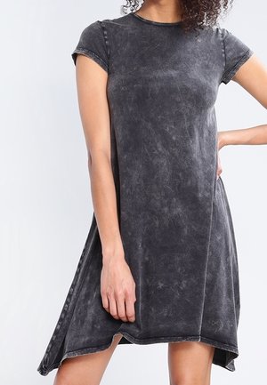 Nainen, joka pukeutuu pestyyn mustaan lyhythihaiseen A-linjaisen mekon, jossa on epäsymmetrinen helma. Hän poseeraa yhdellä kädellä lanteillaan ja toinen käsi roikkuu vartalon sivulla.