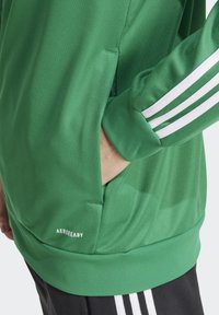 Giacca sportiva verde con una texture liscia, dotata di strisce bianche sulle maniche e un logo AeroReady vicino all'orlo.