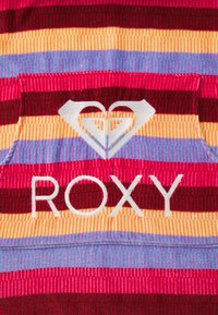 Farebná pruhovaná tkanina s červenými, ružovými, žltými, modrými a tmavohnedými pruhmi. Obsahuje biele logo Roxy a srdcový dizajn. Štruktúrovaný povrch.