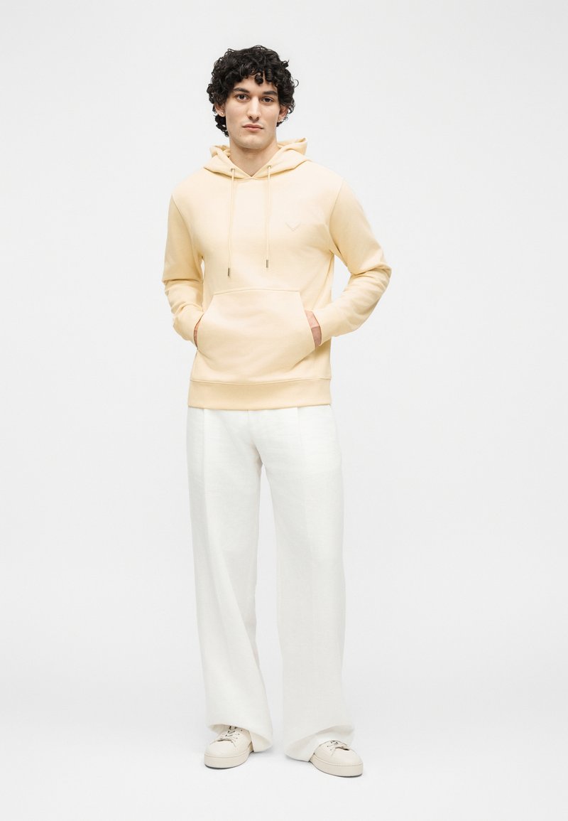 Young person standing with hands in pockets, wearing beige hoodie, white wide-leg pants, and white sneakers against plain light background.