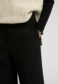 Pantalon noir à la texture lisse, avec des poches latérales dissimulées et une silhouette épurée, accompagné d'un pull en maille épaisse crème.