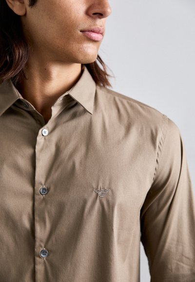 Chemise à manches longues marron clair en tissu lisse, avec un col pointu, un devant à boutons et un logo brodé discret sur la poitrine gauche.