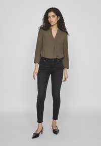 Blusa verde oliva de manga larga con escote en V y textura suave, combinada con jeans negros ajustados y tacones negros de punta.