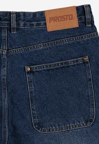 Dżinsy denimowe w ciemnym niebieskim kolorze z skórzaną łatą z napisem "PROSTO". Posiadają tylną kieszeń z złotym nitowym zapięciem i kontrastowym przeszyciem.