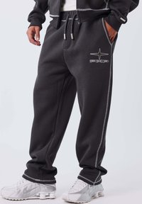 Hombre vistiendo pantalones de chándal gris oscuro con costuras blancas y el logo de PXP, combinados con zapatillas Nike blancas sobre un fondo sencillo.