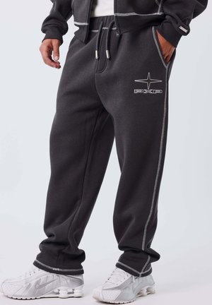 Man in donkere grijze joggingbroek met witte stiksels en PXP-logo, gecombineerd met witte Nike sneakers op een effen achtergrond.