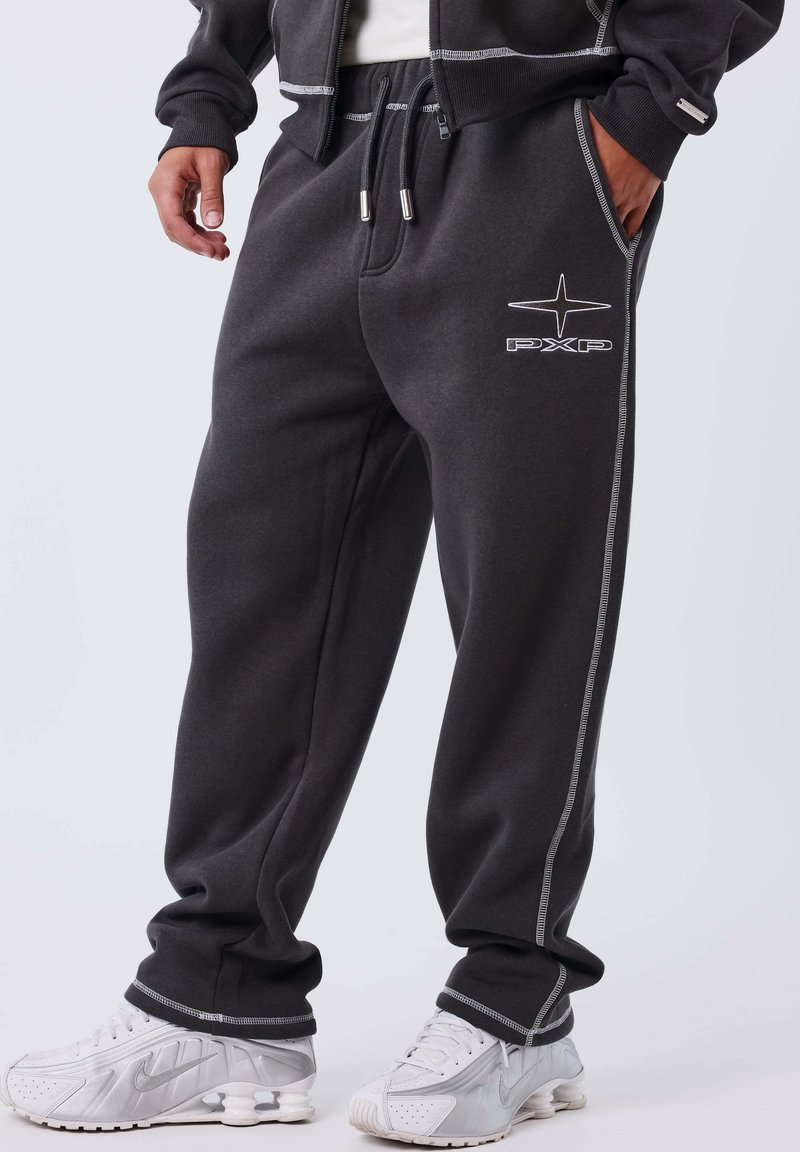 Hombre vistiendo pantalones de chándal gris oscuro con costuras blancas y el logo de PXP, combinados con zapatillas Nike blancas sobre un fondo sencillo.