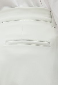 Gros plan sur l’arrière d’un pantalon blanc montrant une seule poche passepoilée et un passant de ceinture, avec une chemise blanche rentrée.