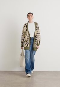 Raksts džemperis zaļā, bēšā un brūnā krāsā ar ribotiem piedurknēm, savienots ar zilām platām biksēm un baltu topu. Aksesuāri ietver bēšu somu.