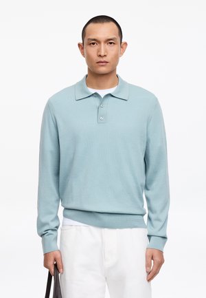 Mężczyzna w jasnoniebieskiej, długorękawowej koszulce polo i białych spodniach, trzymający pasek ciemnobrązowej torby na jednolitym białym tle.