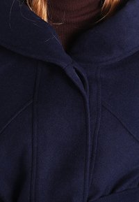 Marinblå ullkappa med huva, med en strukturerad yta, sydda paneldetaljer och en knytning vid halsen.