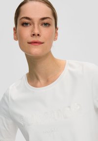 T-shirt blanc à manches courtes avec un texte brodé "NUITS d'ÉTÉ" agrémenté de subtils sequins. Encolure ronde simple et tissu en coton doux.