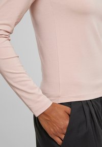 Long-sleeve, light pink shirt in soft fabric, featuring a rounded neckline and fitted sleeves, paired with dark pleated pants.