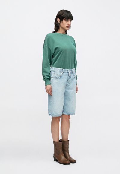 Πράσινο ελαφρύ φούτερ με ριμπ μανσέτες, συνδυασμένο με γαλάζια τζιν βερμούδα και καφέ δερμάτινα μποτάκια αστραγάλου.