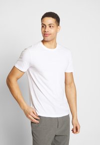 Camiseta blanca de manga corta hecha de algodón, con un clásico cuello redondo, forma ajustada y textura suave, combinada con pantalones grises.