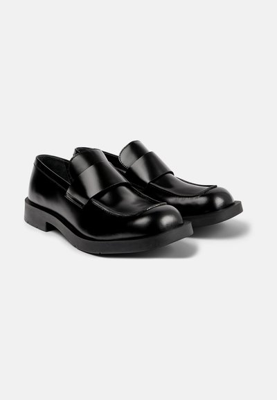 Mocassins en cuir noir avec une finition lisse, présentant de larges lanières sur le dessus, un bout arrondi et une robuste semelle en caoutchouc noir.