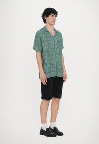 Camiseta verde de manga curta com padrões geométricos, combinada com calções pretos. Modelo usa sapatilhas pretas sem atacadores e meias brancas.