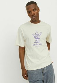 Cream-colored cotton t-shirt with a relaxed fit. Features a purple graphic of a character with raised arms and text "I told you" beneath it.