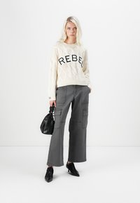 Sweater bege em malha com a inscrição "REBEL" em letras grandes pretas, combinado com calças largas de carga cinzas. Acessórios incluem uma bolsa preta e saltos altos.