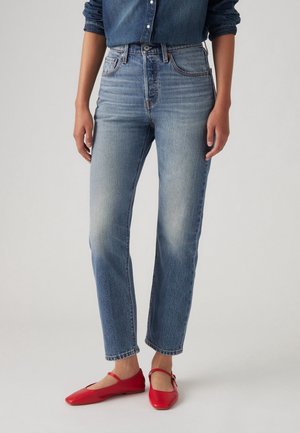 Persona que lleva vaqueros azules de talle alto y pierna recta combinados con zapatos planos rojos estilo Mary Jane y una camisa vaquera azul parcialmente visible.