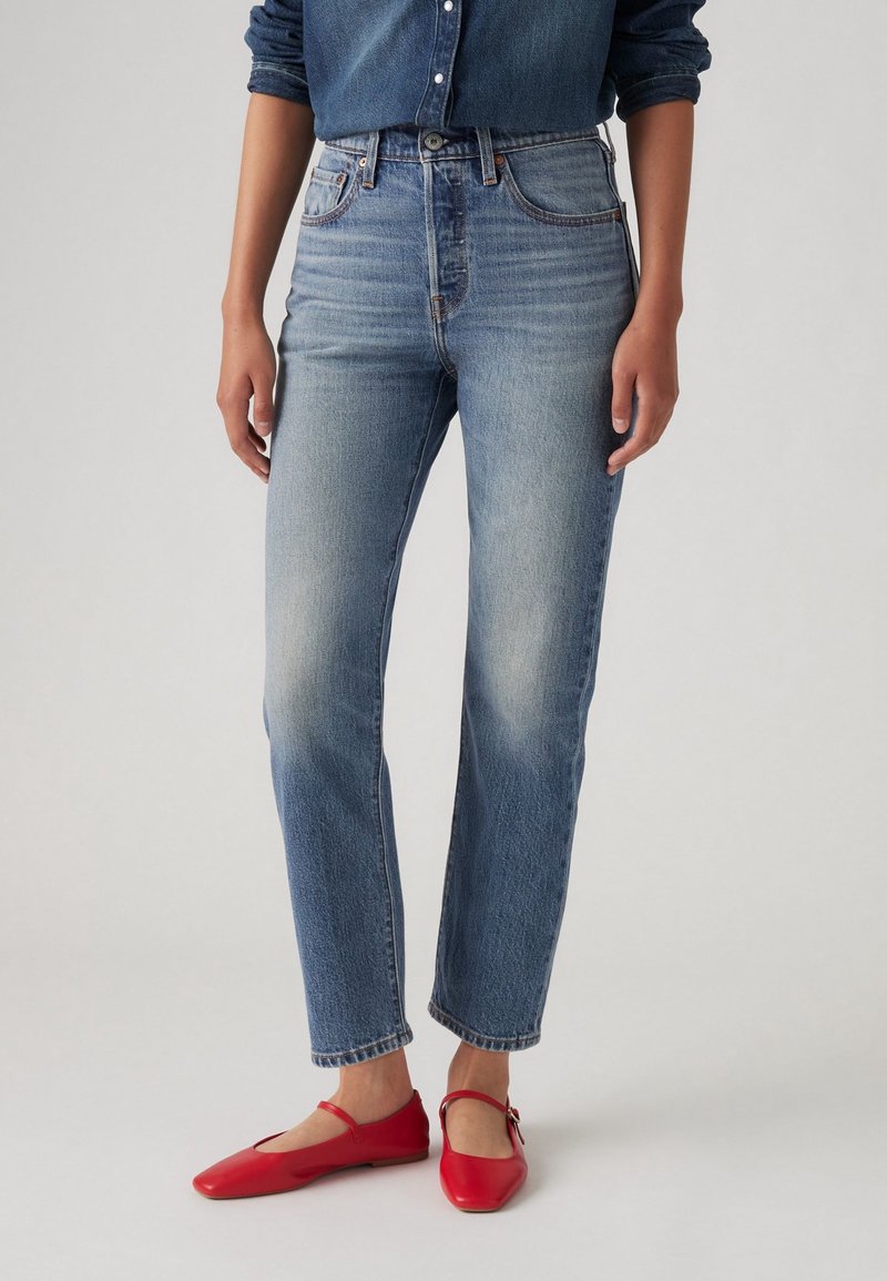 Personne portant un jean bleu taille haute à jambes droites, associé à des ballerines rouges Mary Jane et une chemise en denim bleu partiellement visible.
