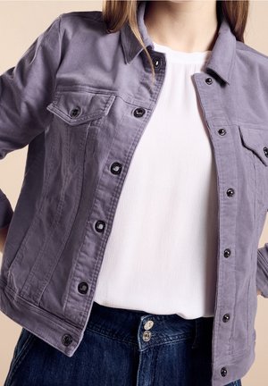 Veste en denim violet avec un col, fermeture à boutons-pression à l'avant et deux poches poitrine. Portée par-dessus un chemisier blanc et une jupe en denim foncé.