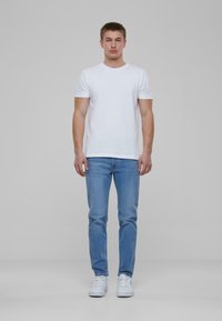 Camiseta de algodón blanca de manga corta combinada con jeans azul claro y zapatillas blancas, de pie sobre un fondo neutro.